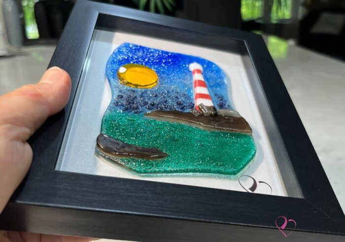 Work in fused glass depicting a lighthouse on a rock overlooking the turquoise sea, framed 5 x 7, Horizon de Percé-Verre collection.