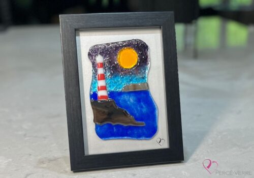 Œuvre en verre fusionné représentant un phare sur un rocher au bord de la mer. Ciel bleu et mauve avec rocher visible à l’horizon. Collection Horizon, cadre 5 x 7.