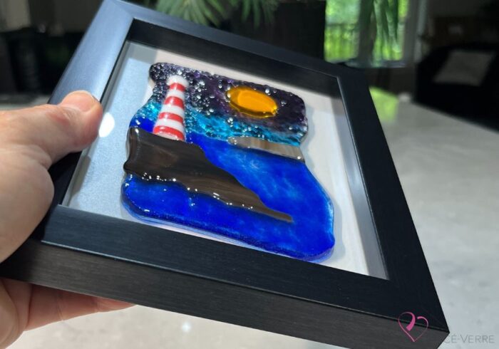Work in fused glass representing a lighthouse on a rock by the sea. Blue and mauve sky with rock visible on the horizon. Horizon Collection, 5 x 7 frame.