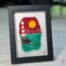 Work in fused glass depicting a lighthouse on a rock by the ocean, under a brilliant red sky. Horizon Collection, 5 x 7 frame.