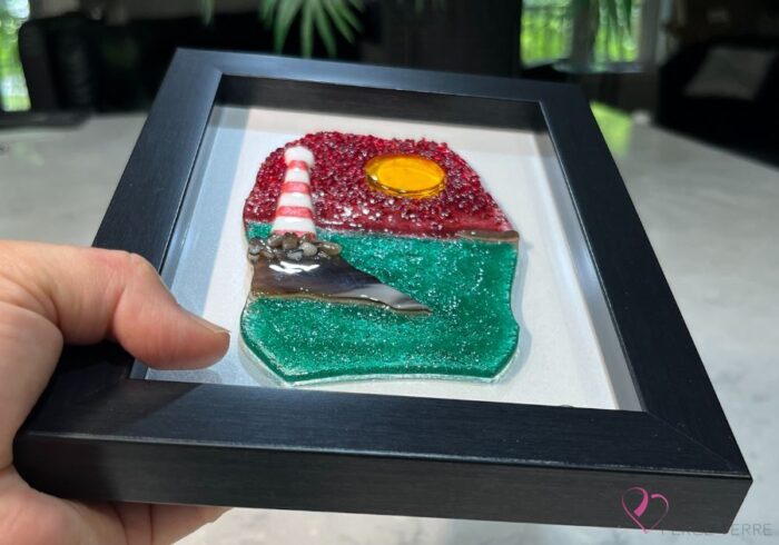 Work in fused glass depicting a lighthouse on a rock by the ocean, under a brilliant red sky. Horizon Collection, 5 x 7 frame.