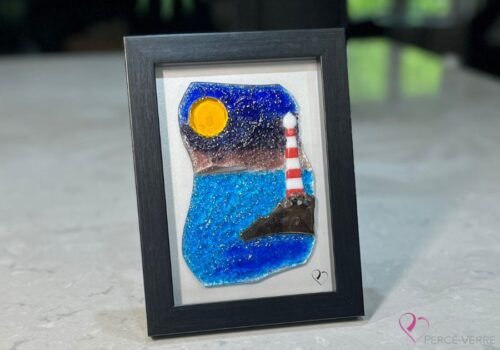 Work in fused glass depicting a lighthouse on the edge of the blue sea, with a touch of mauve in the sky. Horizon Collection, 5 x 7 frame.