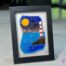 Work in fused glass depicting a lighthouse on the edge of the blue sea, with a touch of mauve in the sky. Horizon Collection, 5 x 7 frame.