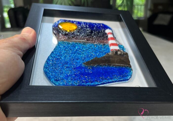 Work in fused glass depicting a lighthouse on the edge of the blue sea, with a touch of mauve in the sky. Horizon Collection, 5 x 7 frame.